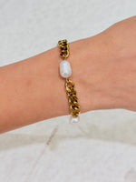 A gold chain bracelet with a central white baroque pearl and a small dangling pearl bead, shown worn on a person's wrist against a light neutral background.